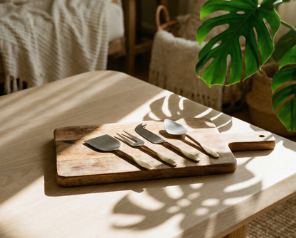 Wooden cutting board with cheese cutlery utensils on a table in a sunlit room with plants