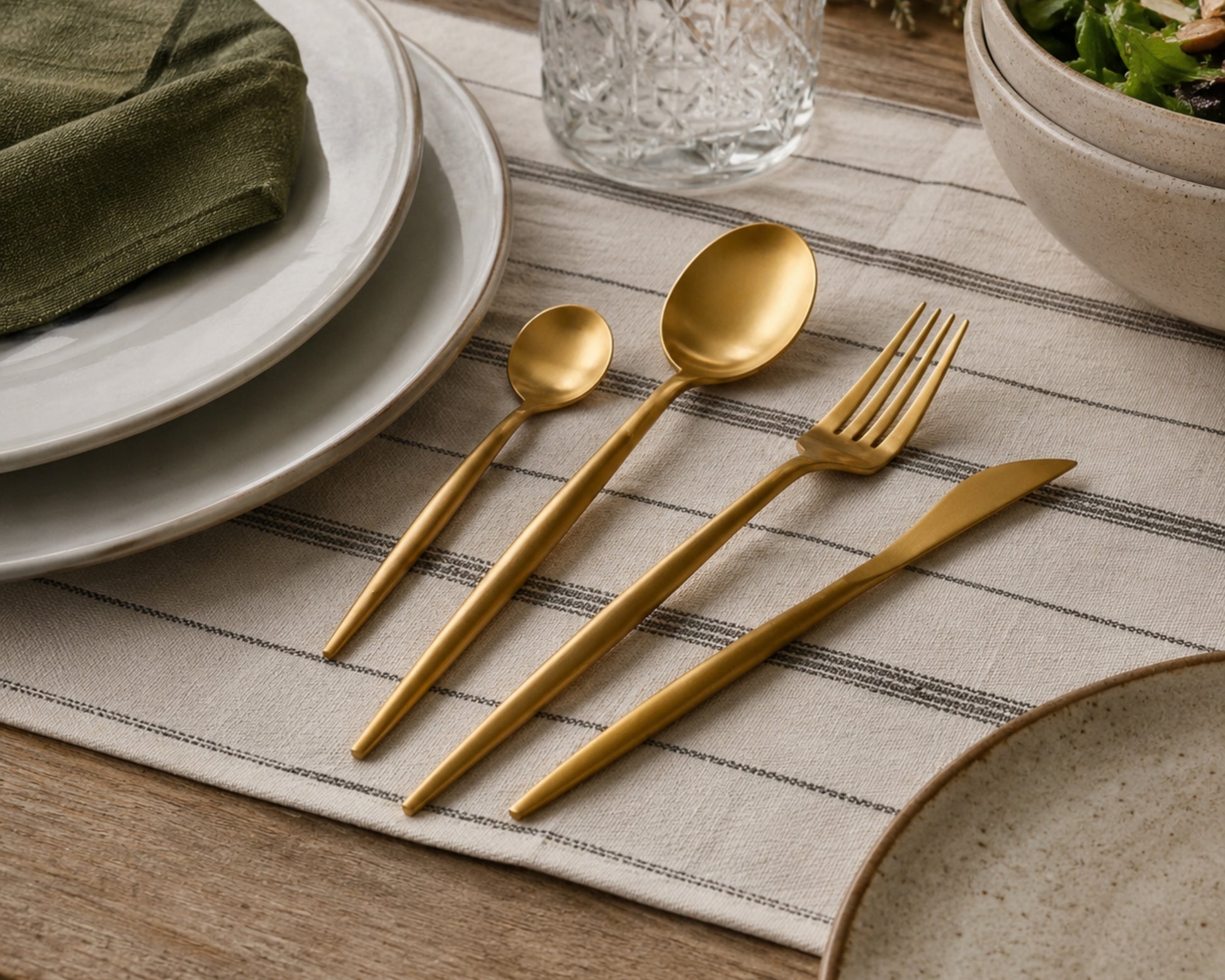 Gold cutlery set on a striped placemat with plates and a bowl in the background.