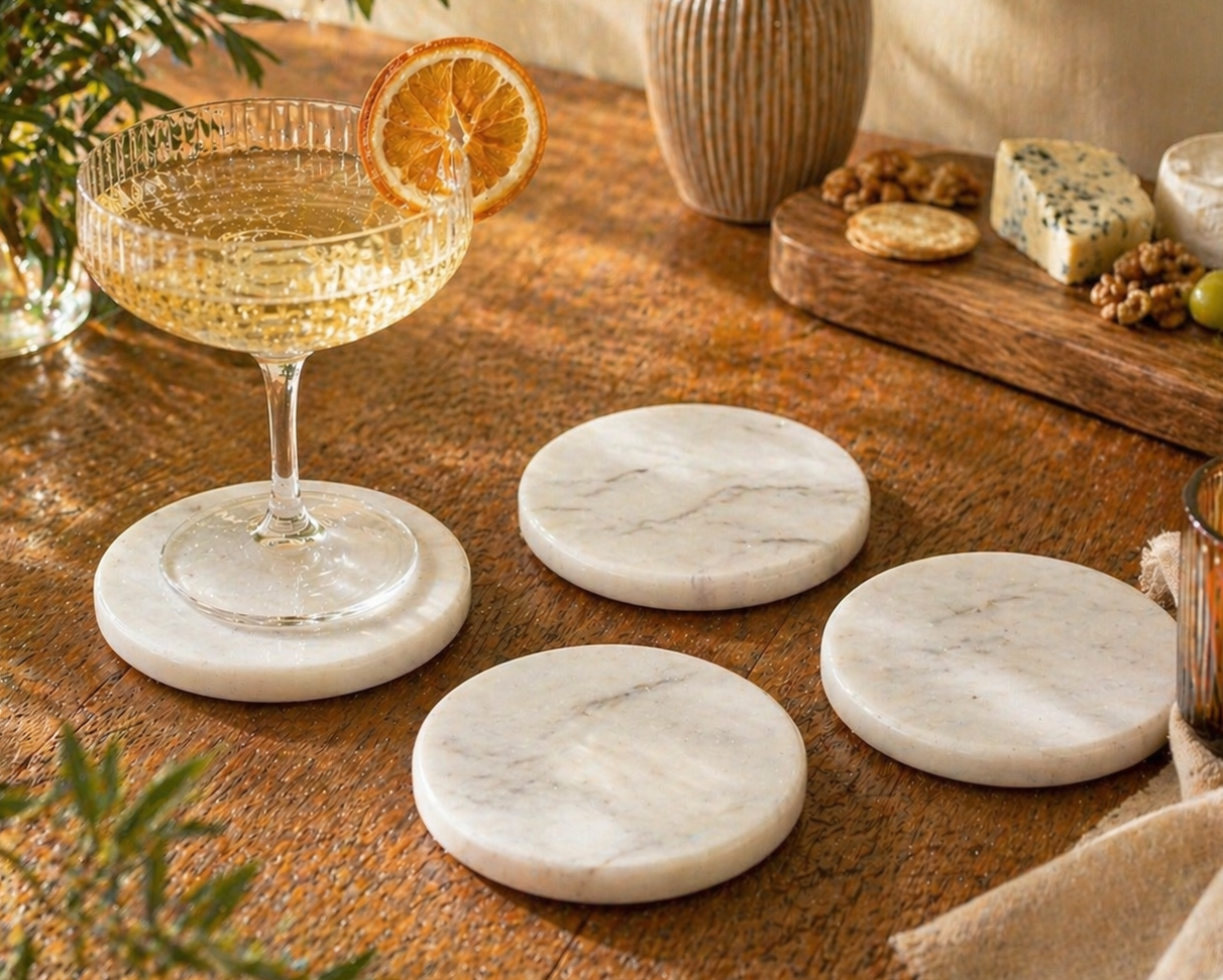 Set of 4 white marble coasters on a wooden table with a glass of white wine, orange slice, and cheese platter.