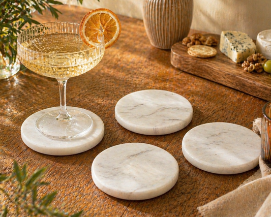 Set of 4 white marble coasters on a wooden table with a glass of white wine, orange slice, and cheese platter.