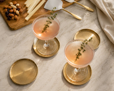 Two cocktails with a sprig of thyme on a marble surface with gold coasters and a charcuterie board.