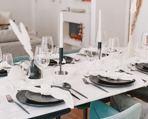 What a Host Tablescapes: Minimal table set up with round black porcelain plates, black stainless steel cutlery, cotton gauze cheesecloth napkins, white cotton gauze table runner, pampas flowers, palm leaf, tapered vanilla white candles, iron black candle holders. Table decoration. Modern tablescape.